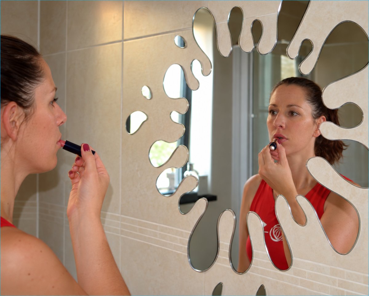 Woman applying lipstick in a splat-shaped Mirrorkool safety mirror fixed to a tiled wall